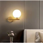 Modern, Electroplated Gold wall sconce featuring an iron horizontal support bar and a large, frosted white glass globe. This elegant fixture uses an E27 bulb and is shown installed over a contemporary bedside table for ambient light.