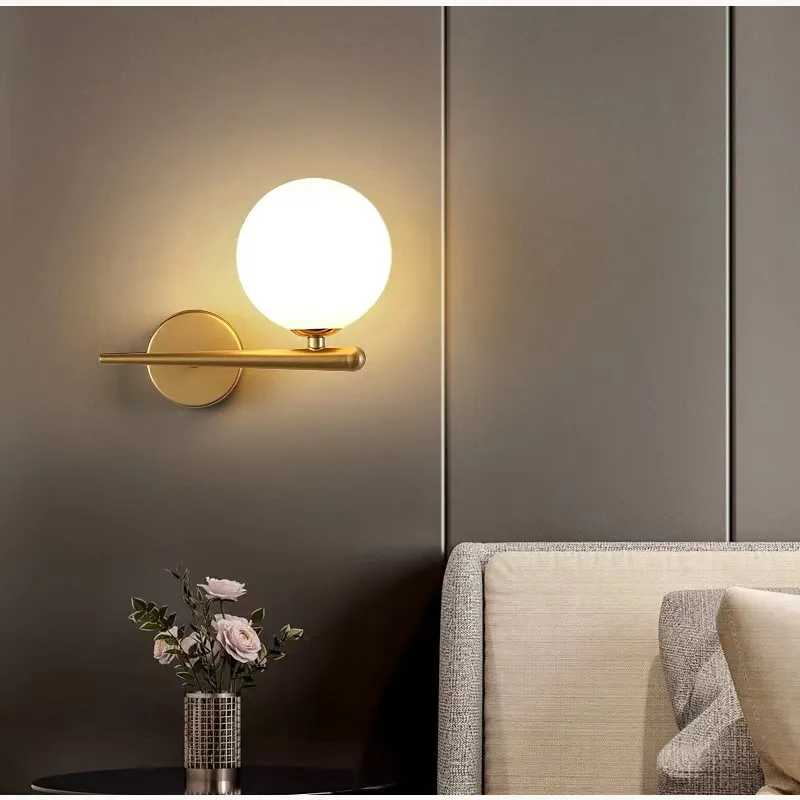 Modern, Electroplated Gold wall sconce featuring an iron horizontal support bar and a large, frosted white glass globe. This elegant fixture uses an E27 bulb and is shown installed over a contemporary bedside table for ambient light.